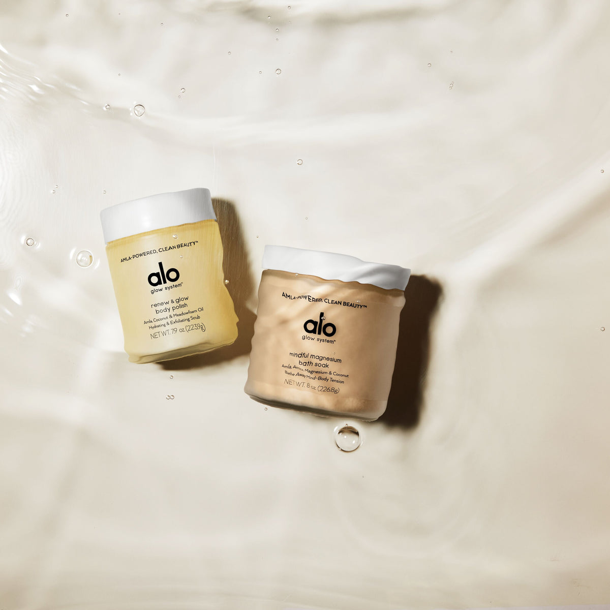 Renew & Glow Body Polish and Mindful Magnesium Bath Soak bottles at the bottom of a beige bathtub under water.