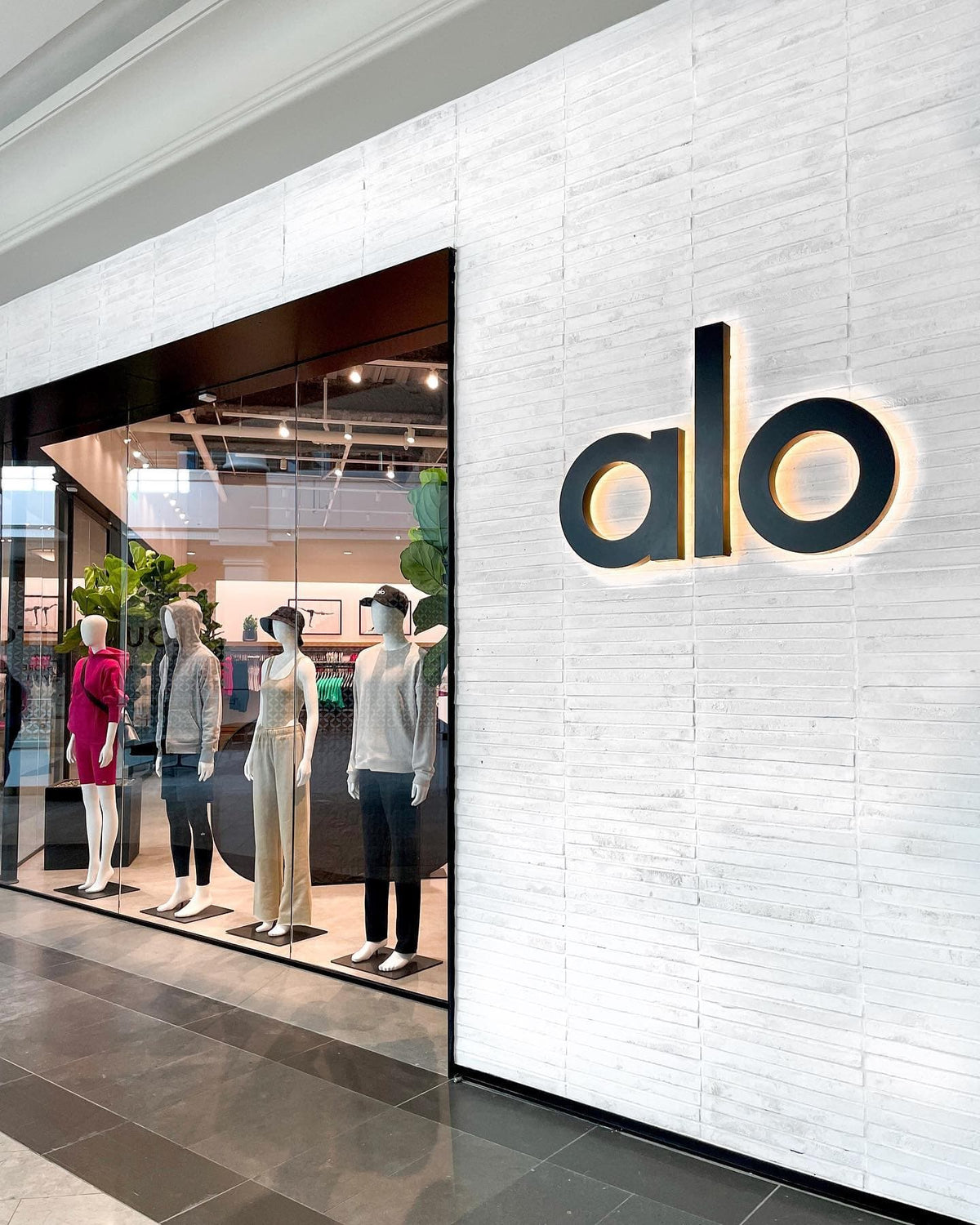 A photo of the Alo Yoga storefront at The Westchester in White Plains New York featuring white brick exterior and black framed floor-to-ceiling display windows lined with mannequins..